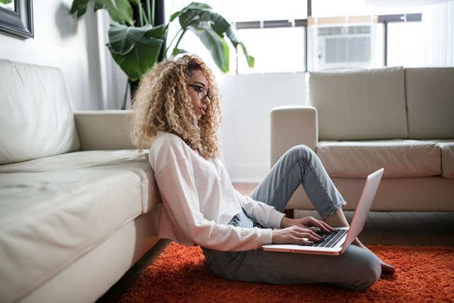 Woman with glasses and curly hair sitting on the floor in a loungeroom with a laptop in her lap